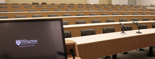 A classroom with six tiered rows of empty brown chairs with no people. The first row has a microphone and Wharton computer.