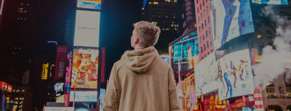 A person in a hooded jacket stands in the middle of a brightly lit urban area, surrounded by large, colorful billboards at night.
