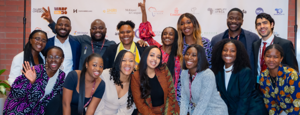 A diverse group of people smiling and posing for a photo in front of a backdrop with various logos, suggesting a professional or networking event.
