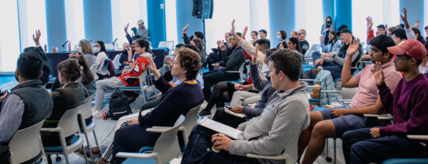 A group of people seated in a classroom-like setting, attentively raising their hands, suggesting participation or answering questions.