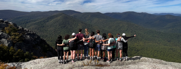 A group of people standing on a rocky cliff with their arms around each other, overlooking a vast, mountainous landscape covered in greenery.