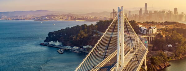 Aerial view of the San Francisco Bay Bridge at sunset, with the city skyline in the background and coastal waters surrounding the bridge.