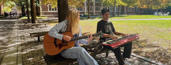 Two people are sitting on a bench outdoors playing musical instruments, one with a guitar and the other with a keyboard, in a sunny park setting.