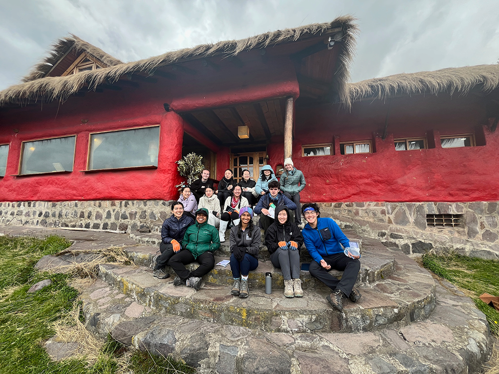 A group of people dressed in outdoor clothing sit on stone steps in front of a red building with a thatched roof and stone foundation. The sky is overcast, and the group appears cheerful and relaxed.