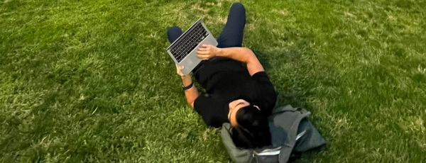 Alex lying on a grass lawn, using a laptop while resting his head on a backpack.